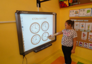 Dziewczynka stoi przy tablicy interaktywnej na której są cztery tarcze zegarów, wskaźnikiem wskazuje na jeden z nich i odczytuje godzinę 9.